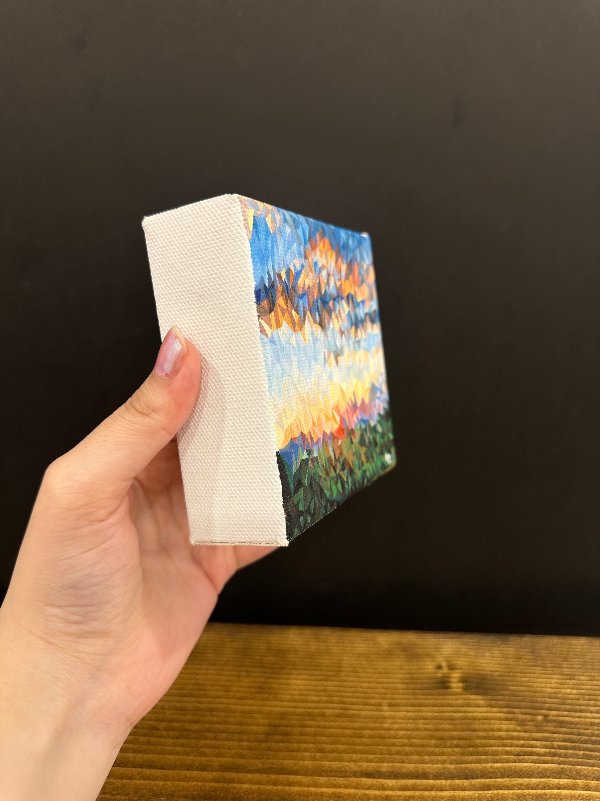 Artist showing the side angle view of 4x4 inches, 10.1x10.1cm oil painting of a bright orange sunrise over a green field, set against a deep blue sky with clouds, rendered in a unique triangles art style, ideal for home decor.